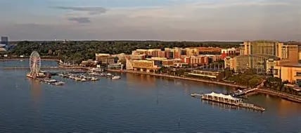Waterfront cityscape with beige buildings along a bay, boats docked at piers, and forested shoreline in background at sunset